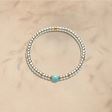 Silver beaded bracelet with gold accents and a single turquoise bead on beige background