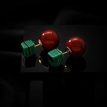 Pair of stud earrings with large glossy red spheres and small green striped cubes on black surface