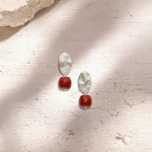 silver oval stud earrings with textured surface and red bead dangle on light textured background