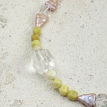 Close-up of handmade beaded jewelry with faceted green stones, clear glass beads, and pinkish irregular pearls on textured surface