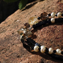 Close-up of a beaded jewelry piece with black, pearl, and amber-colored stones on a textured rock surface