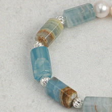 Close-up of a bracelet with rectangular blue-green stones, silver beads, and a pearl on a gray background