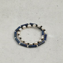 Two beaded bracelets with white pearls, black and silver beads, and blue cylindrical stones on gray surface