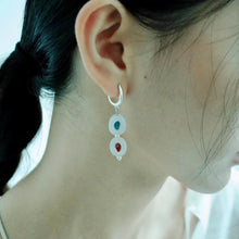 Close-up of a woman's ear wearing silver hoop earrings with hanging white discs and blue and red stones