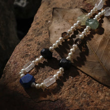 Pearl and gemstone necklace with blue, black, green, and clear beads on dry leaf and stone