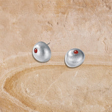 Silver stud earrings with a small red gemstone on a textured beige surface