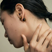 Close-up of a woman wearing brown spherical stud earrings with black hair tied back, hand touching neck