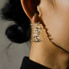 Close-up of a woman's ear wearing modern silver abstract dangle earrings against dark hair and neutral background
