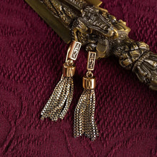 Gold tassel earrings with decorative rectangular plaques on textured burgundy fabric