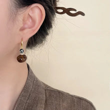Close-up of a woman's ear wearing a dangle earring with speckled and brown beads, brown hair clip, beige background