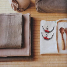 Folded beige and pink linens, cotton pouch with metal rings, and wooden spoons on woven mat