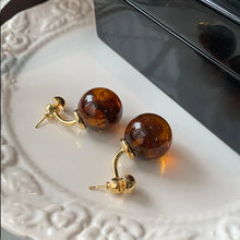 Pair of gold stud earrings with round amber-colored resin balls on a white decorative plate