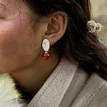 Close-up of a woman's ear wearing a silver fingerprint stud earring with a red bead drop, dark hair, and beige coat