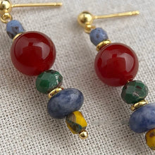 Close-up of colorful beaded earrings with red, green, blue, and yellow stones on beige fabric