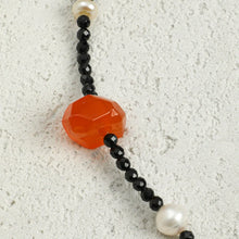 Close-up of faceted orange gemstone bead with small black beads and white pearls on textured light surface