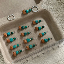 Handcrafted turquoise and brown beaded jewelry charms with silver loops displayed on a beige tray next to a ruler