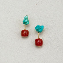 Pair of stylish earrings with turquoise blue irregular top stones and red barrel-shaped beads on light textured background