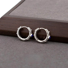 Silver hoop earrings with intricate swirl patterns and small blue stone accents on gray fabric surface