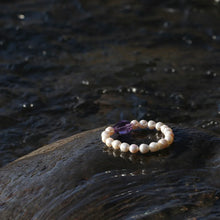 Pearl bracelet with purple gemstone resting on wet rock by flowing water