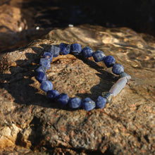 Close-up of a blue sodalite gemstone and white pearl bracelet on a textured rock surface under sunlight