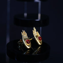 Gold hand-shaped earrings with red stone accents on black glossy display stand