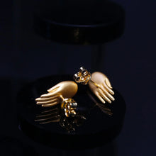 Gold hand-shaped stud earrings on a black reflective surface with dark background