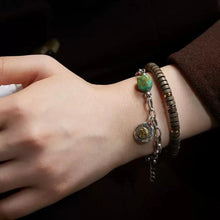 Wrist with two bracelets, one silver chain with green stone and charm, and one brown beaded bracelet