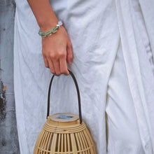 Hand wearing green beaded bracelet holding bamboo woven handbag over white fabric dress