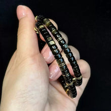 Hand holding two black and gold beaded bracelets against a black background