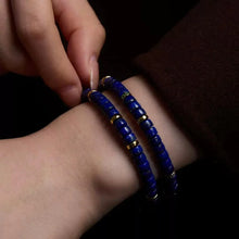 Close-up of hand wearing two blue beaded bracelets with gold accents on wrist against dark background