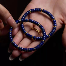 Close-up of hands holding blue and gold beaded bracelets with a gold ring on finger