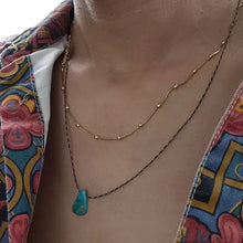 Close-up of layered gold bead necklace and black chain necklace with turquoise pendant on person wearing colorful patterned shirt