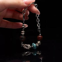 Hand holding a silver chain bracelet with mixed beads in turquoise, brown, and metallic colors on black background