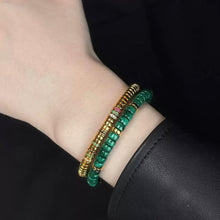 Wrist with two beaded bracelets, one gold with colored stones and one green, against a black sleeve