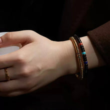 Hand wearing a gold ring and two bracelets, one gold and one black with colorful beads, on wrist