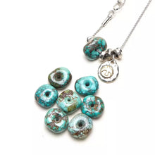 Turquoise donut shaped beads and silver chain necklace with sun and bird charm on white background
