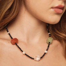 Close-up of woman's neck wearing a beaded necklace with black beads, pearls, and large red, green, and clear stones