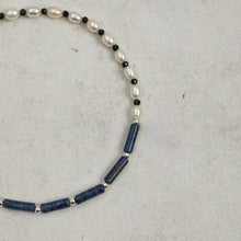 Close-up of a beaded bracelet with white pearls, black beads, blue cylindrical stones, and silver spacer beads on gray background